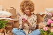 © WHstudio Leushin N - Positive curly haired woman texts messages via smartphone happy to get nice offer of buying new house gathers personal belongings in cardboard boxes prepares for moving. Real estate service.