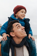 Wall Mural - Handsome young smiling father carrying on shoulders his little cute son while spending fun time together on beautiful beach