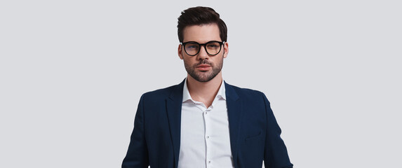 Wall Mural - Handsome young man in eyeglasses looking at camera while standing against grey background