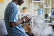 Side view of young black man in t-shirt standing in front of camera in living room and texting in smartphone while smoking e-cigarette