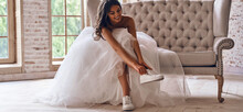 Modern Young Woman In Wedding Dress Putting On Sports Shoes And Smiling While Sitting On The Couch