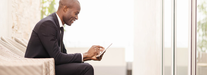 Wall Mural - Checking his schedule on the go. Side view of cheerful young African businessman working on digital tablet while sitting on the chair