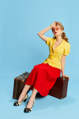 Wall Mural - Feeling tired. Beautiful young girl in yellow blouse and red skirt sitting on vintage suitcases against blue studio background. Concept of retro fashion, beauty, travelling, 50s, 60s. Pin-up style