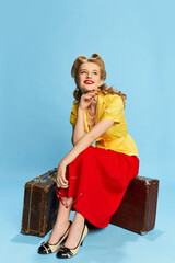 Wall Mural - Dreamy happy look. Beautiful young girl in yellow blouse and red skirt sitting on suitcase against blue studio background. Concept of retro fashion, beauty, travelling, 50s, 60s. Pin-up style