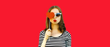 Portrait Of Beautiful Young Woman Covering Her Eyes With Red Sweet Heart Shaped Lollipop And Blowing Her Lips On Background