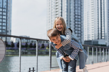 Poster - happy preteen boy in denim outfit piggybacking girl on river embankment.