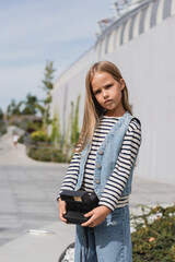 Wall Mural - preteen girl in denim vest and striped long sleeve shirt holding vintage camera near mall building.