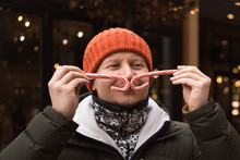 Man In Hat And Winter Clothes Makes Mustache From Candy Canes, Posing For Photo At Christmas Market