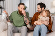 Disgusted gay parent holding diaper near husband and baby daughter in living room.