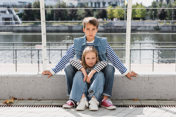 Wall Mural - stylish kids in denim vests and jeans sitting near metallic fence on riverside.