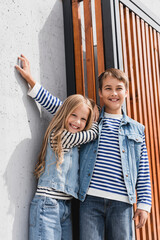 Wall Mural - portrait of happy children in striped long sleeve shirts and denim vests looking at camera near building.