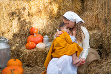Mom Holds In Her Arms A Little Daughter With Bare Feet, Wrapped In A Yellow Cloth, Sitting On Burlap In Stacks Of Straw And Pumpkins And Gently Hugs
