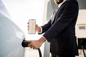 Crop of man in black suit plugging power cord and showing mobile screen with app of charging status of electric car battery. Technology and innovation concept.