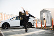 © sofiko14 - Charming young woman in business suit dancing joyfully outdoors while her luxury electro car charging on EV station. Enjoyment of free time with modern eco friendly transport.