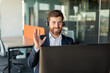 © Prostock-studio - Happy businessman in headset working on computer and waving his hand to webcamera, having video call in office interior