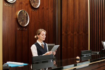 Young blond female receptionist in uniform looking at tablet screen while standing by counter in lounge of modern hotel and networking