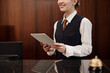 © pressmaster - Close-up of young woman in uniform of receptionist holding tablet while standing by workplace in front of camera and waiting for guests