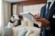 © pressmaster - Close-up of young male manager of hotel using digital tablet while looking through list of booked guests with chambermaid on background
