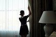 Back view of young chambermaid in uniform opening heavy night curtains in hotel room while standing in front of large window in the morning