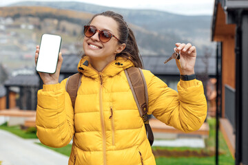 Young female in yellow down jacket shows application on smartphone near modular cabin houses