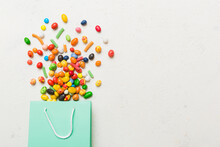 Shopping Paper Gift Bag In Corner Full Of Assorted Traditional Candies Falling Out On Colored Background With Copy Space. Happy Holidays Sale Concept