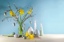 Seasonal Holiday Decoration With Candles, A Bouquet Of Daffodils And Spring Branches In A Glass Vase And Artificial Gray Easter Eggs On A Light Wooden Board Against A Blue Wall, Copy Space