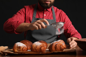 Canvas Print - Male baker sprinkling croissants with sugar powder at table on dark background, closeup