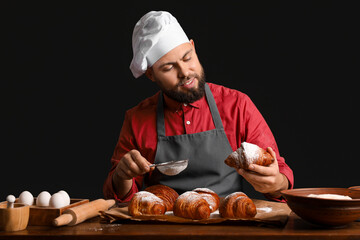 Canvas Print - Male baker sprinkling croissants with sugar powder at table on dark background