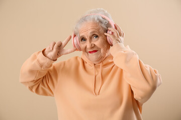 Wall Mural - Senior woman in headphones showing victory gesture on beige background