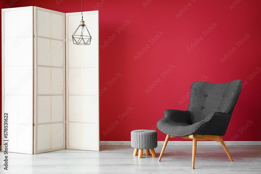 Stylish grey armchair, folding screen, pouf and lamp near red wall