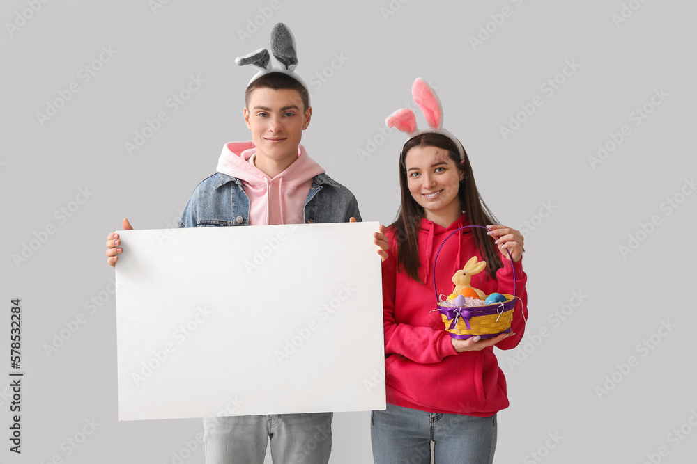 Happy young couple in bunny ears with Easter eggs and blank poster on grey background