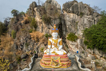 The White Buddha Statue With Rock Mountain In The Background At Tham Champathong Monastery, Ratchaburi, Thailand