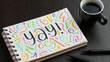 YAY! black lettering with colorful motifs in notebook with cup of coffee and pens on black wooden desk