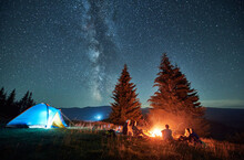 Night Camping In Mountains Under Starry Sky. Group Of People Tourists Having A Rest Near Campsite, Burning Campfire And Illuminated Tent. Concept Of Tourism, Hiking And Adventure.