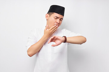 Sleepy young Asian Muslim man in Arabic clothes looking wristwatch, covering his mouth with hand while yawning isolated on white background. People religious Islamic lifestyle concept