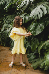 Little girl in a yellow dress in a botanical garden. a child stands near the leaves of Monstera.