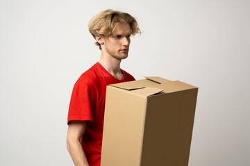 Sticker - Delivery man holding a cardboard boxes and smiling while standing against white background. Courier holding a cardboard box in front of himself.