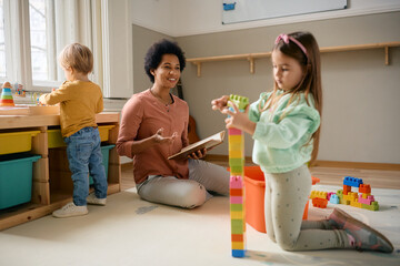 Wall Mural - Small girl stacking toy blocks while playing with her African American teacher at preschool.