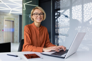 Portrait of happy and successful female programmer inside office at workplace, worker smiling and looking at camera with laptop blonde businesswoman is satisfied with results of achievements at work