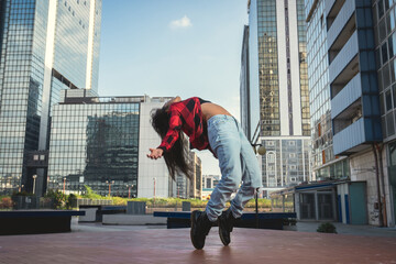 Photo shoot of stylish caucasian hip hop dancer posing on her toes with red shirt, jeans and black boots shoes and top with a urban town as background. City shooting of moving model bending the back