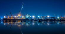 Shipyard Dry Dock Maintenance And Repair Container Ship Transport And Oil Ship Tanker, Crane Work And Commercial Port Reflection In Water, At Night Over Lighting In Sea Long Exposure Blue Color Tone,