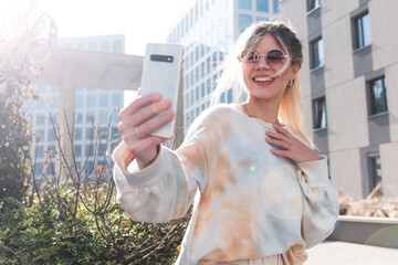 A young beautiful girl blogger takes selfies among the cityscape on a sunny day.