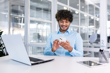 young indian programmer inside modern office sitting at workplace, man using phone, testing online a