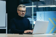 © Liubomir - Serious mature gray-haired businessman working inside modern office, man thoughtfully typing on laptop at workplace.
