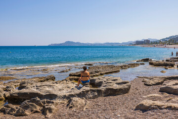 Beautiful seascape view. Woman sitting on rocks near water and looking horizon. Blue water surface merging with cloudless sky. Greece