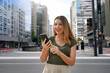 © zigres - Cheerful Brazilian girl watching funny video on phone standing on street at daytime. Brown-haired with toothy smile wears green t-shirt. Social media addiction concept.