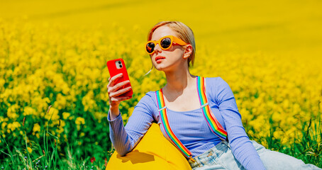 Sticker - Beautiful female in rainbow LGBT suspenders for pants and mobile phone is sitting in bean chair in rapeseed field