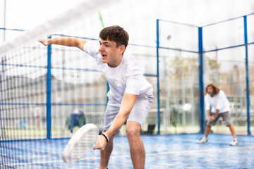 Wall Mural - Portrait of emotional young sporty guy playing paddle tennis with friends on open court. View through net