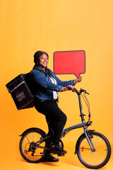 Wall Mural - Pizzeria courier holding red speech bubble advertising takeaway service while riding bicycle preparing to deliver takeout order to customers. Courier standing in studio with yellow background