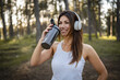 One woman happy caucasian female standing in the forest or park in woods with supplement shaker and headphones during training in nature taking a brake water drink and hydration real people copy space